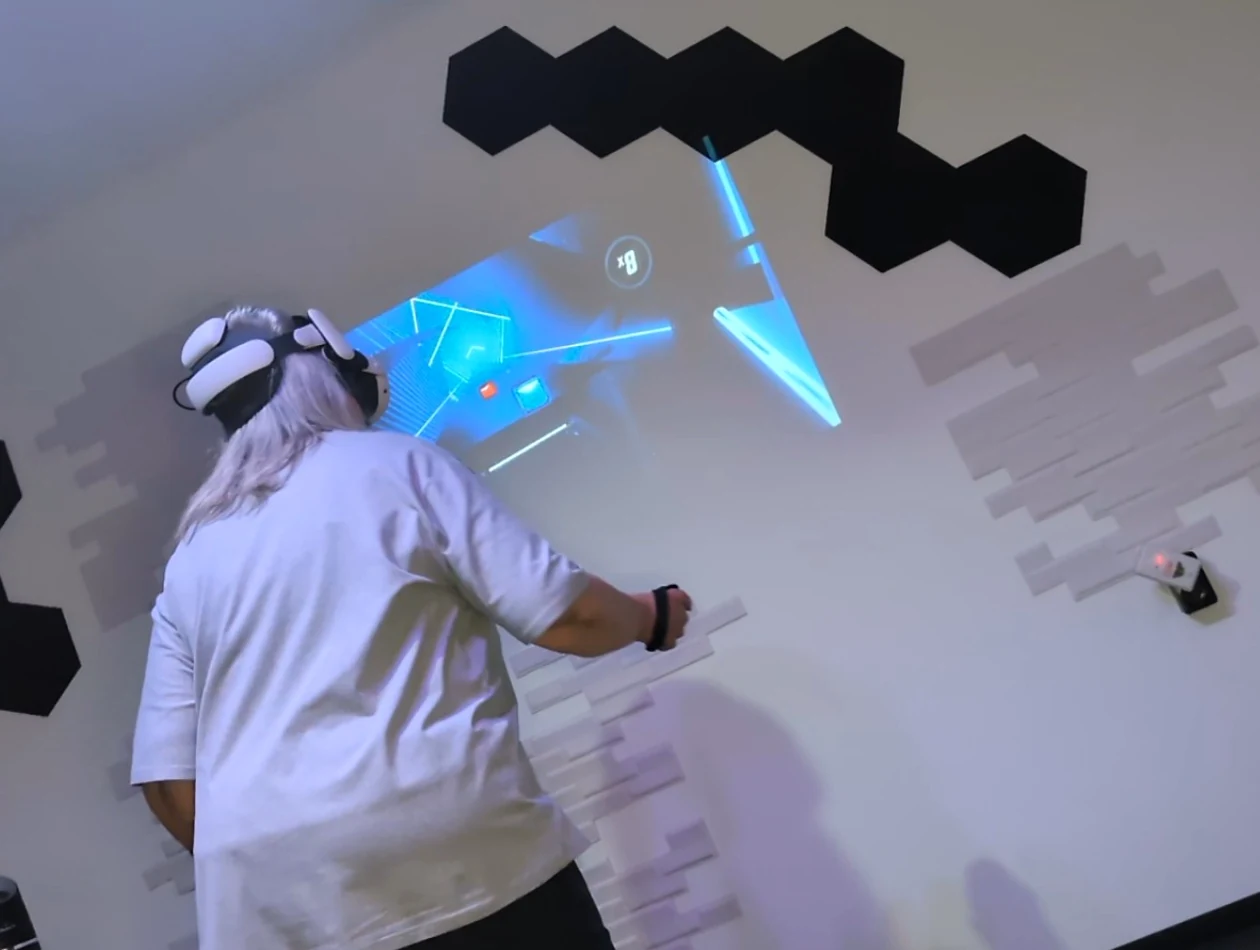 Osoba w goglach VR wyciąga rękę, a na ścianie widać projekcję odpowiadającą temu, co dzieje się w wirtualnym świecie.